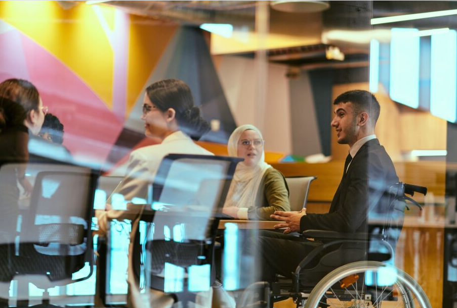  In a modern glass startup office, a wheelchair-bound director leads a successful meeting with colleagues, embodying inclusivity and teamwork.