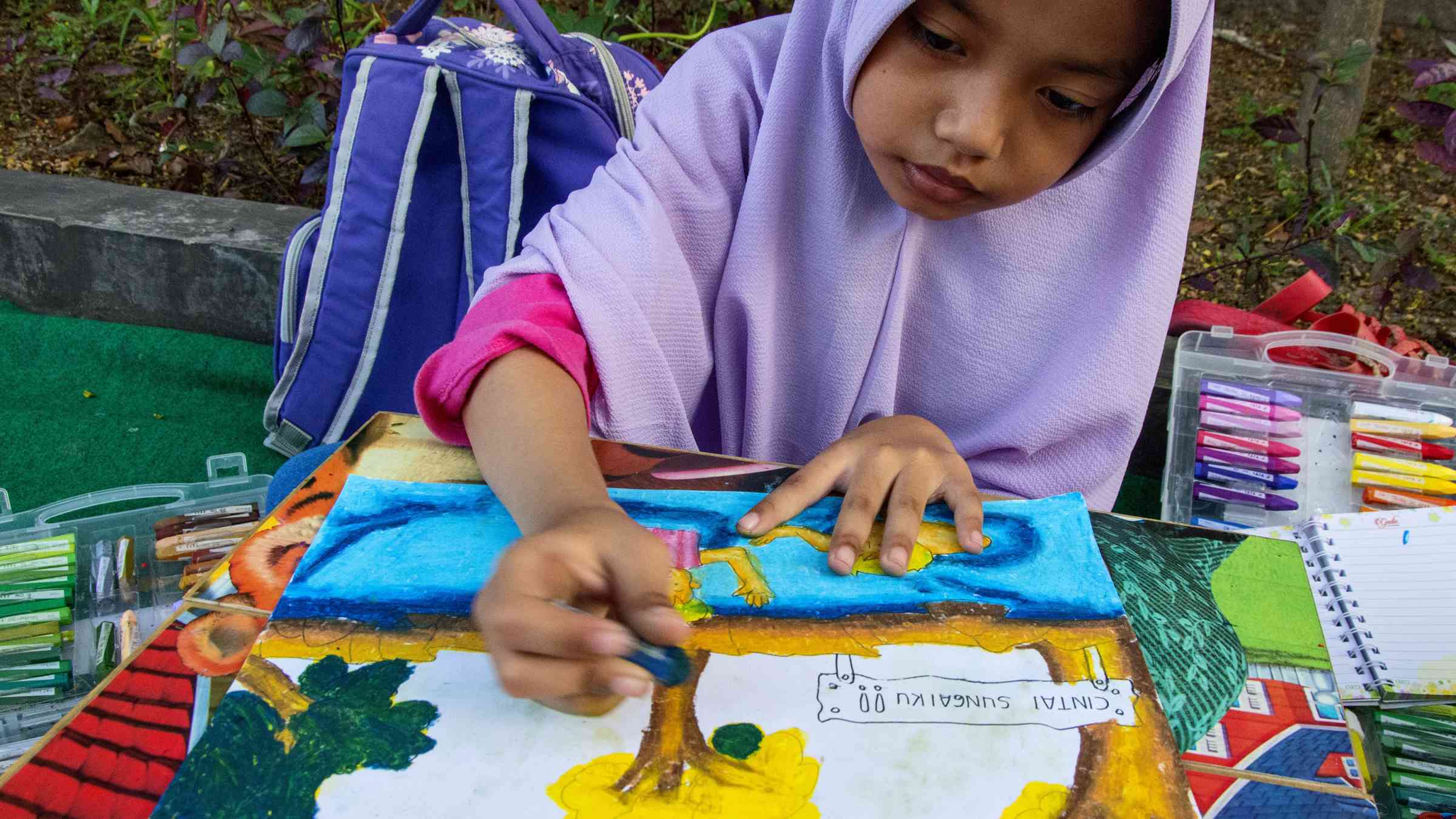 A girl completing a painting showing trees, people and a sign saying “Cinta Lingkungan”