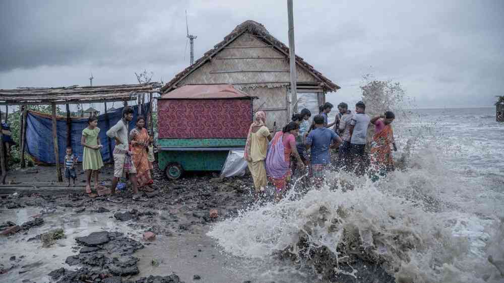 A group of people by a wooden house on the coast as a tidal wave breaks on the land
